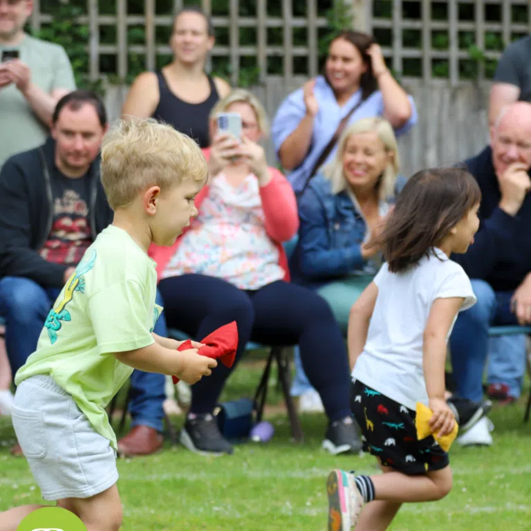 Two kids run on a grassy field, each holding colored cloths, while spectators sit and cheer in the background.