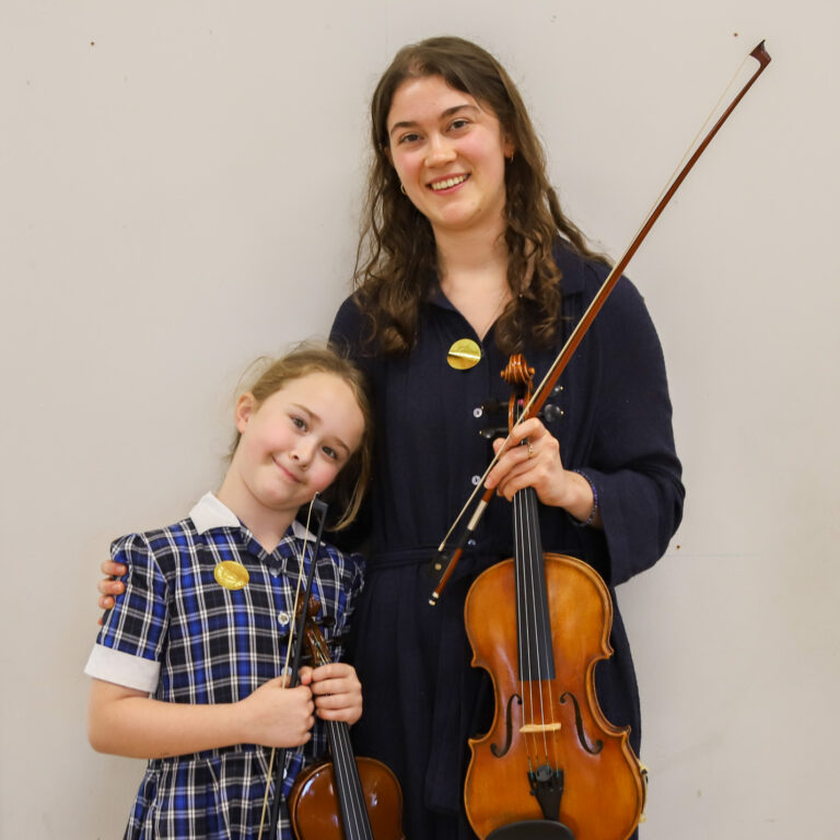Two young violinists pose together, each holding a violin and bow, smiling against a plain light wall.
