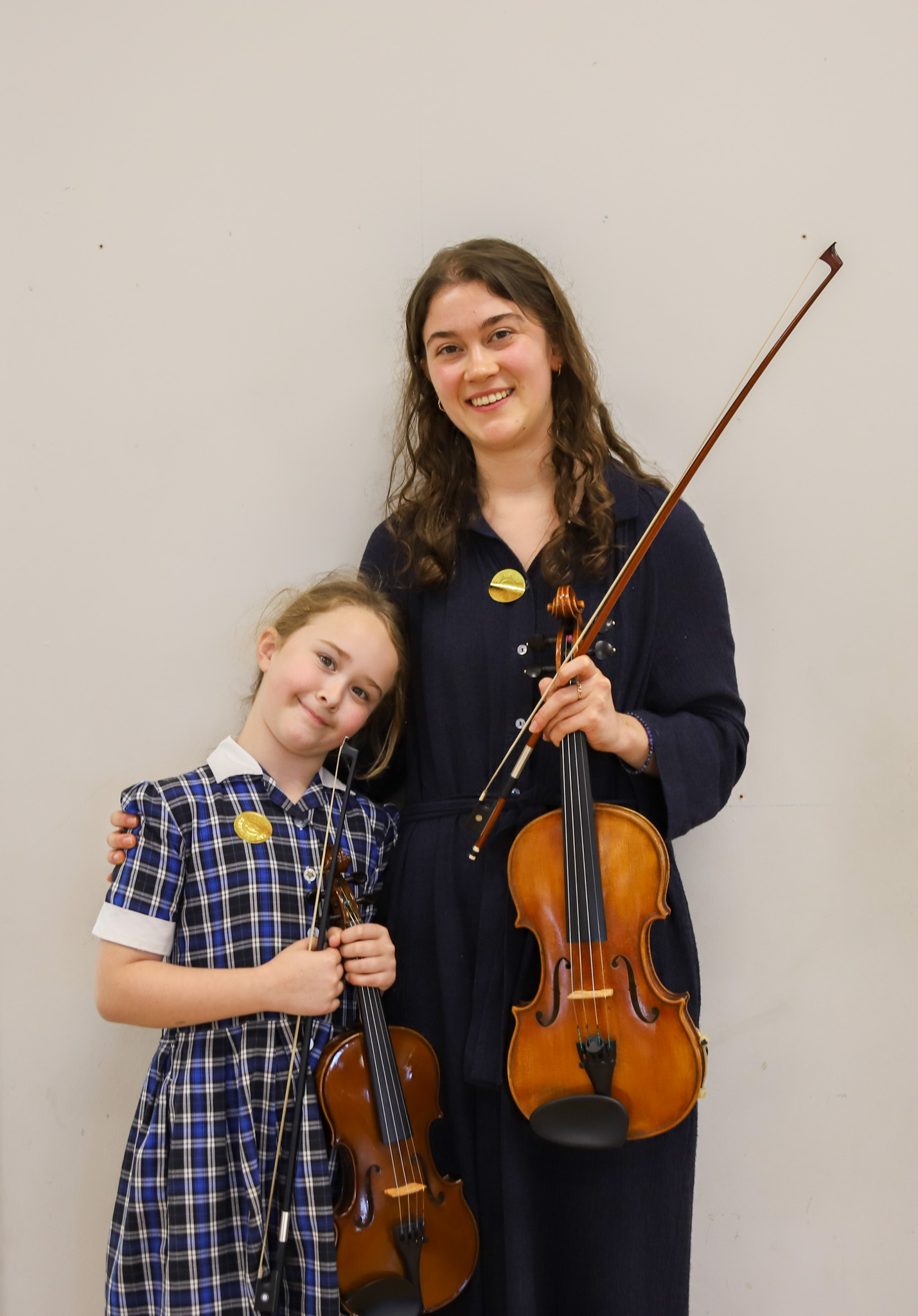Two young violinists pose together, each holding a violin and bow, smiling against a plain light wall.
