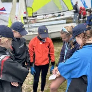 Group of young sailors in hats and jackets forming a circle on a grassy shore, studying a map before a sailing session, with colorful sailboats in the background.