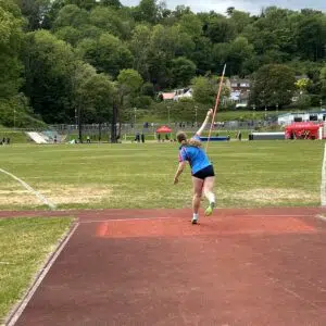 Young female athlete in a blue shirt and black shorts throws a javelin on a red track, with a grassy field and trees in the background.