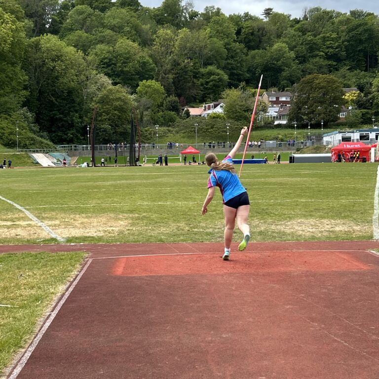 Young female athlete in a blue shirt and black shorts throws a javelin on a red track, with a grassy field and trees in the background.