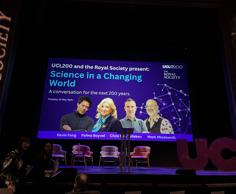 Stage with a large screen reading 'Science in a Changing World' and four panelists pictured, podium center, chairs on stage, audience in front.