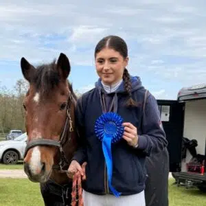 Young rider stands with a brown horse, holding a blue rosette ribbon earned in a horse show outdoors. alsa field and trailer in background.
