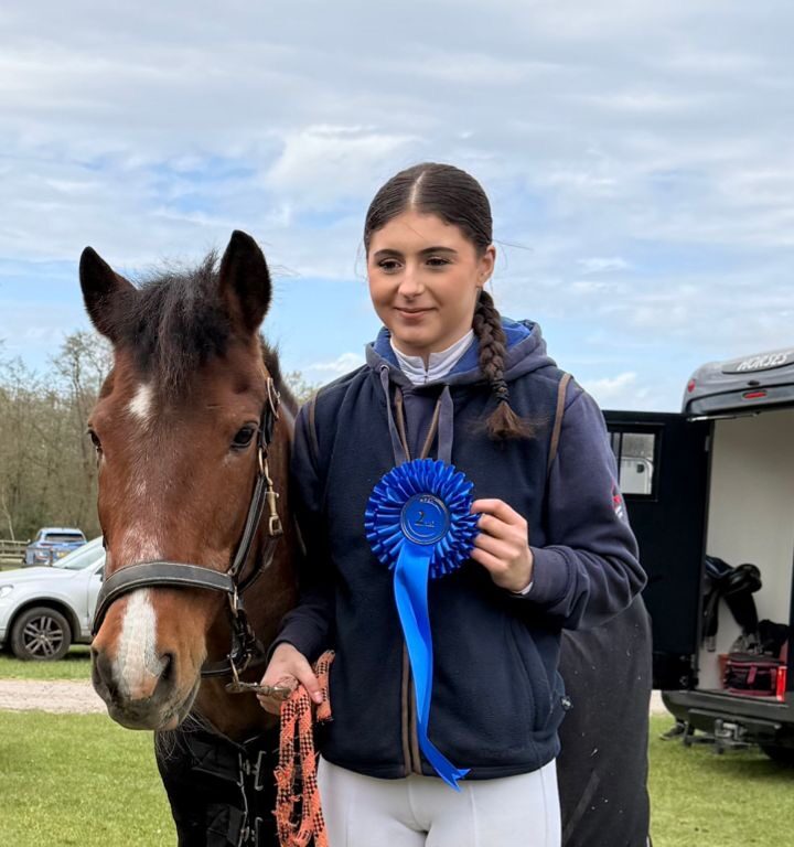 Young rider stands with a brown horse, holding a blue rosette ribbon earned in a horse show outdoors. alsa field and trailer in background.