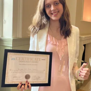 Young woman in a pink ruffled blouse and white blazer holding a framed certificate and a glass award, smiling at the camera in an indoor setting.