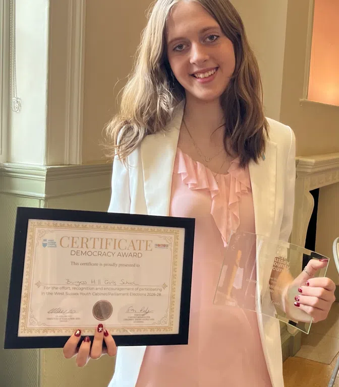 Young woman in a pink ruffled blouse and white blazer holding a framed certificate and a glass award, smiling at the camera in an indoor setting.