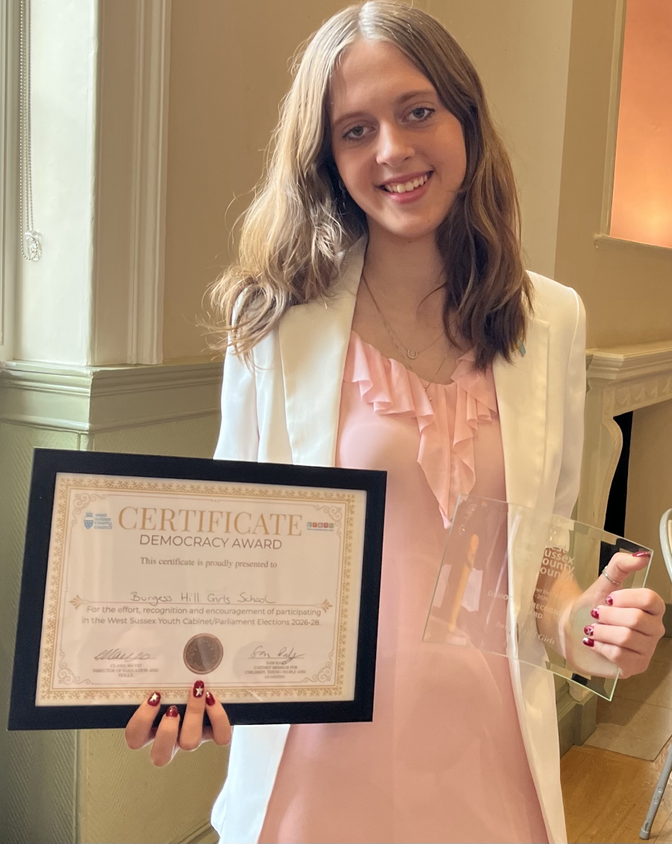 Young woman in a pink ruffled blouse and white blazer holding a framed certificate and a glass award, smiling at the camera in an indoor setting.