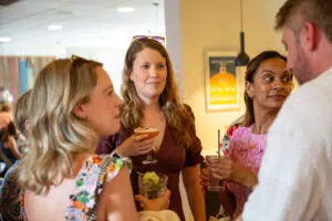 Group of friends chatting at a bar or party, holding cocktails and smiling at each other indoors.