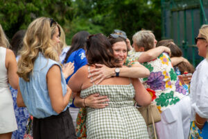 Group of people hugging at an outdoor gathering, smiling faces, greenery in the background.