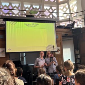 Two students present on a library stage in front of a bright lime-green screen reading Burgess Hill Girls 'Pourquoi ?' while classmates watch.