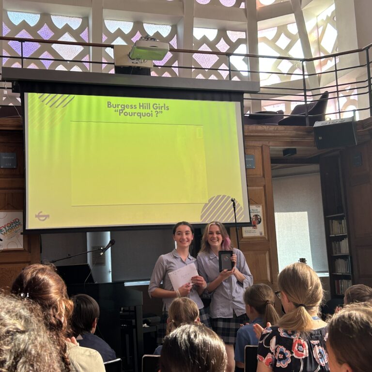 Two students present on a library stage in front of a bright lime-green screen reading Burgess Hill Girls 'Pourquoi ?' while classmates watch.