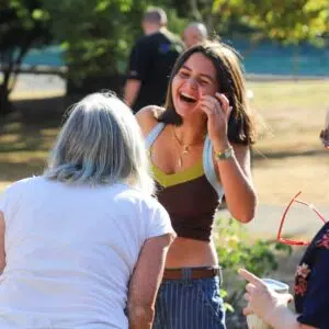 Young woman laughing and talking with two others outdoors in a sunny park setting, one with white hair facing away in the foreground.