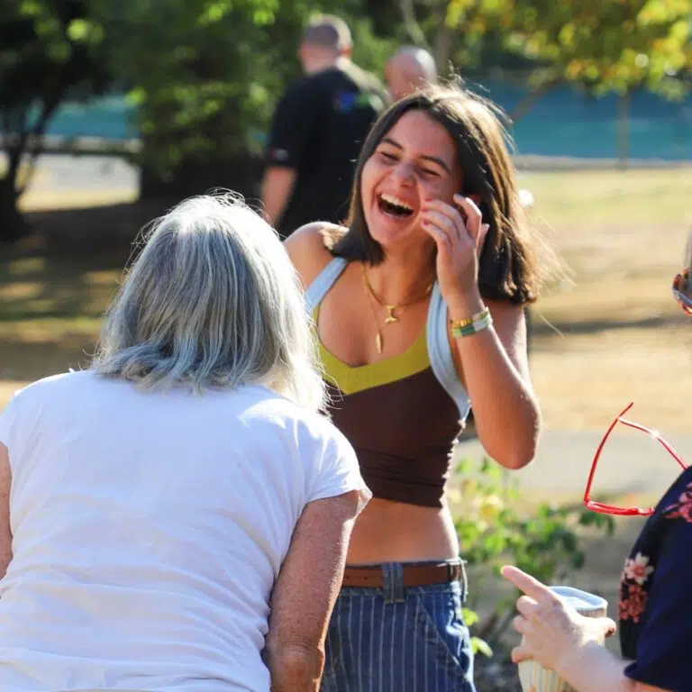 Young woman laughing and talking with two others outdoors in a sunny park setting, one with white hair facing away in the foreground.