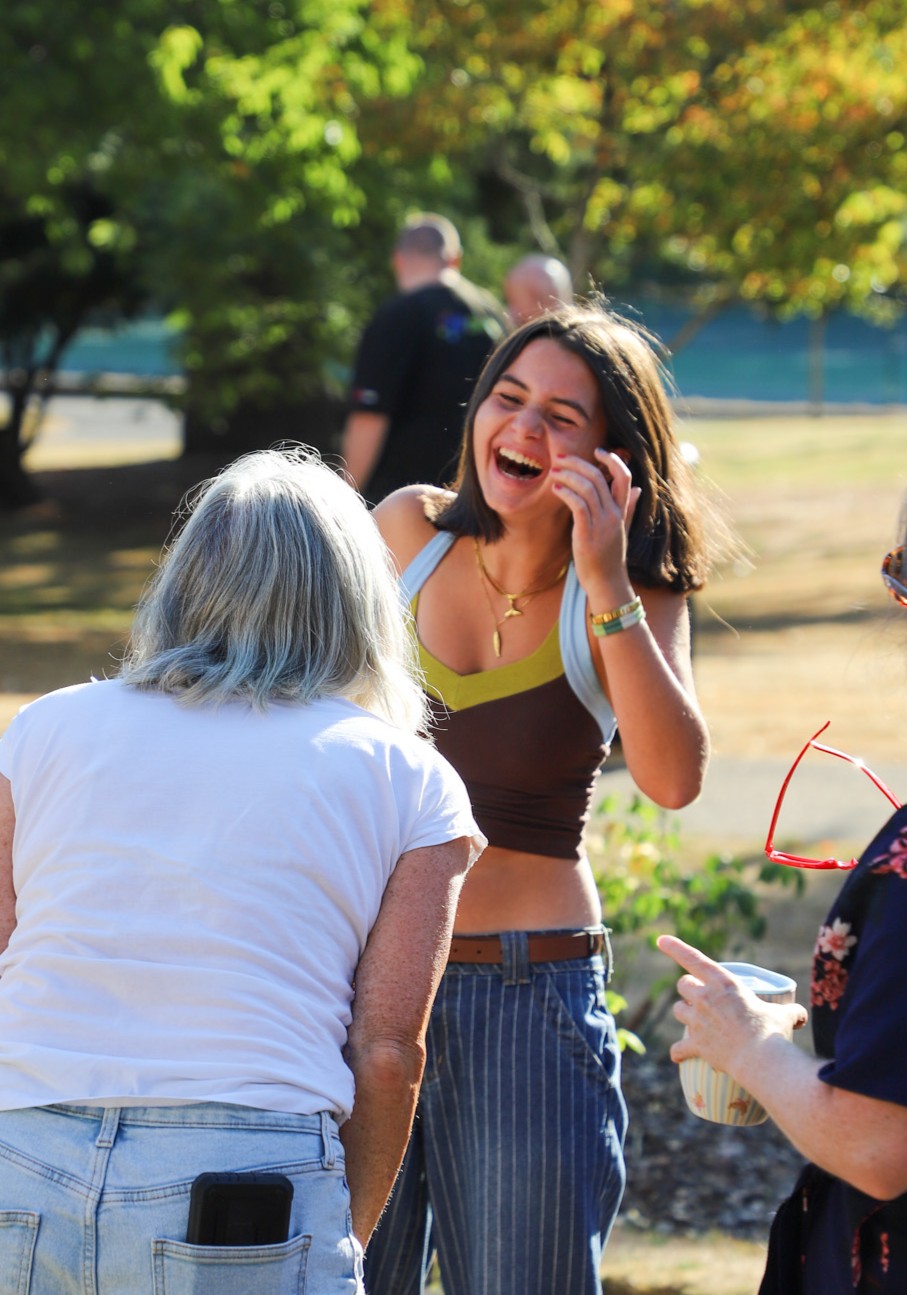Young woman laughing and talking with two others outdoors in a sunny park setting, one with white hair facing away in the foreground.