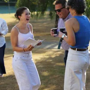 Young woman in white tank top and pants laughs while holding papers, talking with two people outdoors on a sunny day.