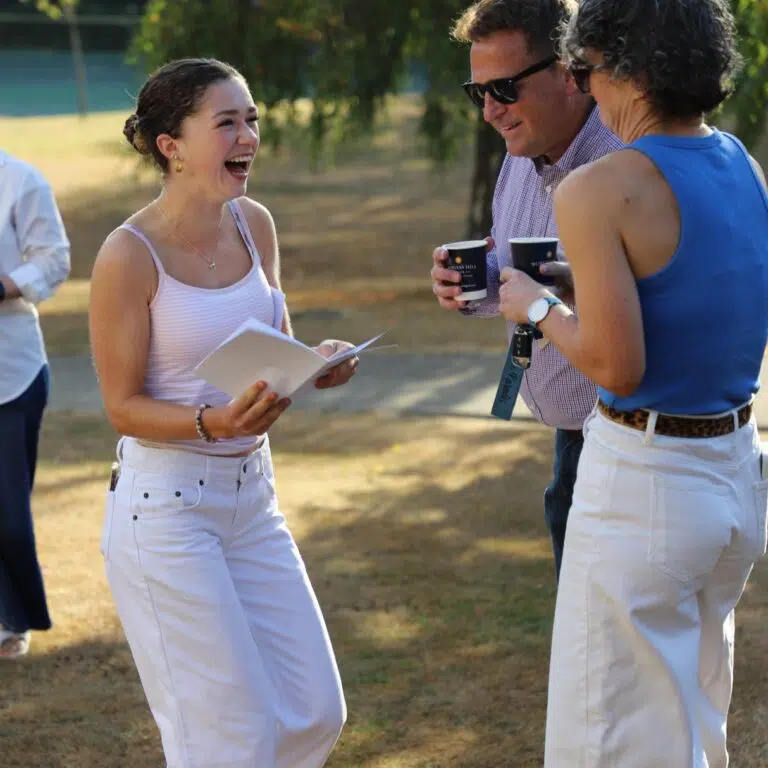 Young woman in white tank top and pants laughs while holding papers, talking with two people outdoors on a sunny day.