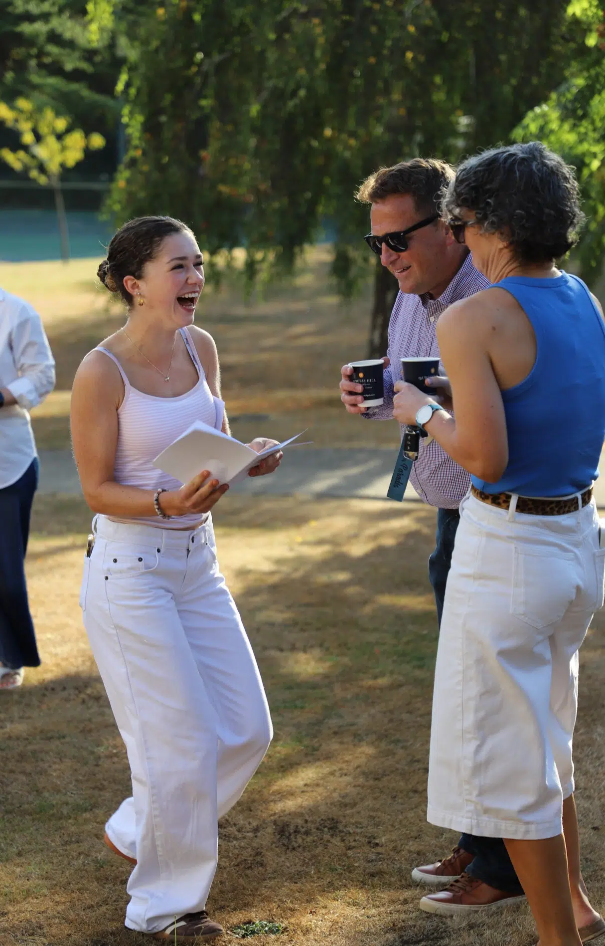 Young woman in white tank top and pants laughs while holding papers, talking with two people outdoors on a sunny day.