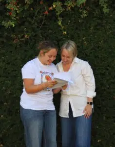 Two women stand outside in front of a green hedge, smiling as they read papers together.