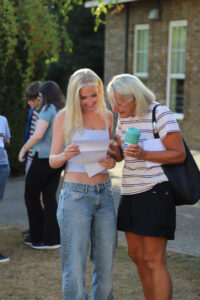 Two women outdoors, smiling, looking at papers together in front of a brick building.