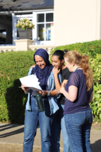 Three women stand outside smiling while looking at papers; one wears a navy hijab, others in casual jeans and dark top.
